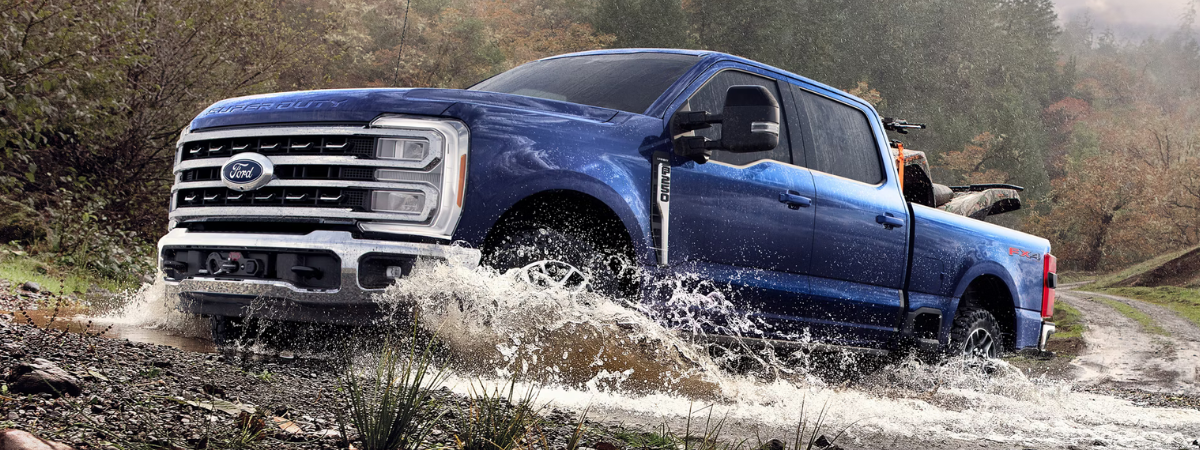 A blue Ford F-250 Super Duty truck with an ATV in its bed, splashing through a muddy puddle on a dirt road.
