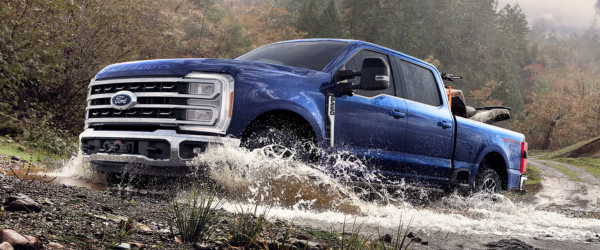 A blue Ford F-250 Super Duty truck with an ATV in its bed, splashing through a muddy puddle on a dirt road.